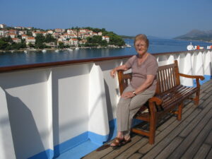 Elderly woman wearing soft colours sitting on a bench on a boat with sea and sky in background.