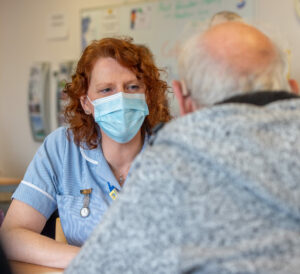 An elderly man, sat at a table, talking to a St Barnabas Health Care Support Worker.