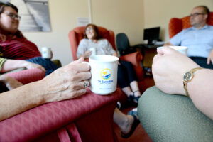 Person holding white mug with St Barnabas Hospice logo on, part of a circle of people sitting in chairs.