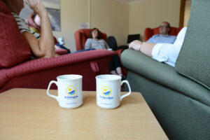 Two white mugs with St Barnabas Hospice logo on wooden table with chairs and people in background.