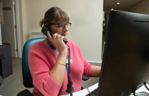 Woman wearing pink top speaking on the phone in front of computer monitor.