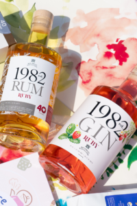 Two bottles with white labels. On the right, Est.1982 Ruby Gin with pink drink, and on the left Est.1982 Ruby Rum with yellow liquid.
