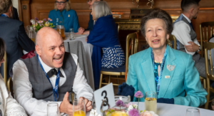 Man wearing shirt and waistcoat talking whilst sitting at a table with HRH Princess Anne, wearing a blue blazer.