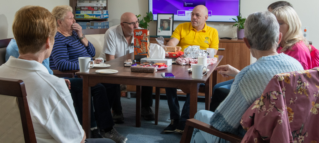 Man in yellow polo shirt, speaking with people at a table that has cups, biscuits and games on it.