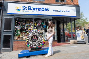 Young brunette woman standing by heart-shaped sculpture in front of St Barnabas Hospice charity shop.