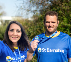A man and a woman wearing a St Barnabas running tops. The Man is holding a medal, stood in front of green foliage.