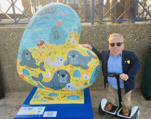 Actor Warwick Davis, patron of St Barnabas, on Segway next to heart shaped sculpture with paintings of seals.