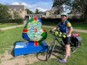 A women, wearing cycling clothing and equipment, on a bike, in front of a Heart shaped culture. The Heart shaped sculpture is decorated with a Lincolnshire flag.