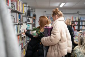 Two women in winter coats browsing a bookshelf inside The Warehouse