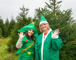 Two woman dressed in elf and tree Christmas costumes stood in front of real Christmas trees