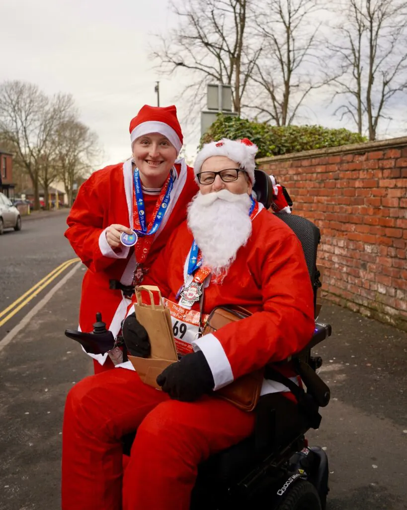 Geoff in a Santa Suit in his wheelchair, with his niece stood next to him also in a Santa Suit, both smiling at the camera after finishing the event